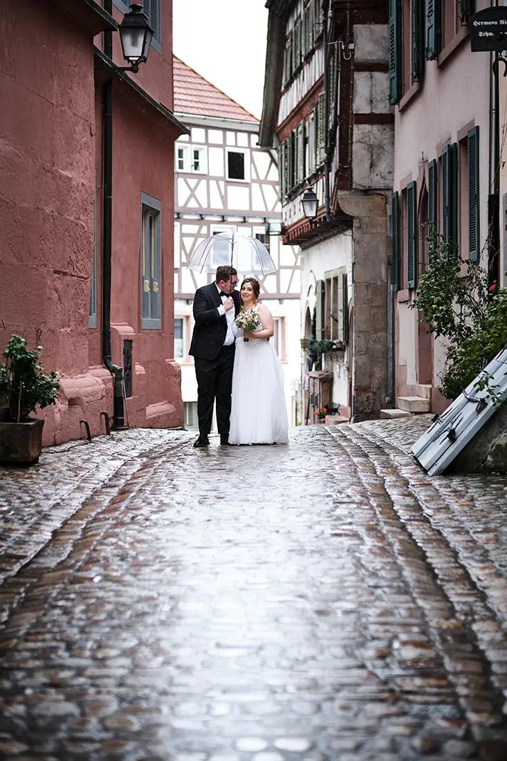 Romantisches Hochzeitsfoto in der Altstadt von Gernsbach bei Regen – das Brautpaar unter einem transparenten Regenschirm auf nassem Kopfsteinpflaster.